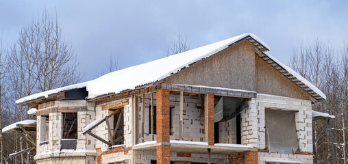 Unfinished home roofing metal tiles construction is covered with snow against cloudy sky. House...