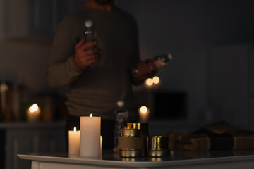cropped view of blurred man holding bottled water near table with stock of canned food near candles...