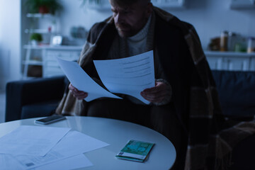 man under warm blanket looking at payment bills near money and smartphone during electricity outage.