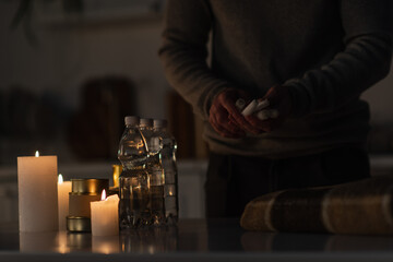 partial view of man holding candles near reserve of bottled water and canned food on table.