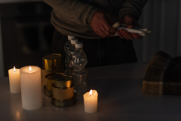 partial view of man with candles near table with warm blanket and reserve of water and canned food.