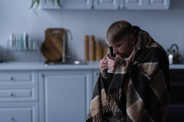 frozen man in warm blanket drinking hot beverage during energy blackout at home.