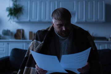 displeased man looking at invoices while sitting in kitchen under blanket during power shutdown.