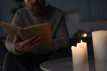 cropped view of man reading book near candles burning in dark kitchen during power outage.