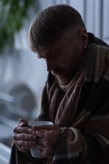 frozen man wrapped in warm blanket holding cup of hot tea during power shutdown at home.