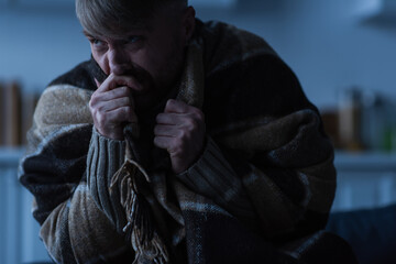 stressed man holding hand near face while sitting under warm blanket and looking away in twilight.