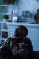 frozen man wrapped in warm blanket sitting in blurred kitchen with closed eyes.