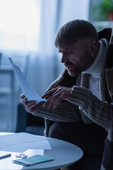 frozen man in warm sweater and blanket looking at payment bills during electricity shutdown.