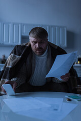 worried man under warm blanket holding smartphone and invoices in twilight at home.