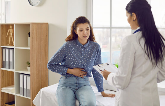 Female patient with abdominal pain undergoing medical examination by doctor for indigestion. Young woman is talking to doctor and holding her stomach while sitting on medical couch in doctor's office.
