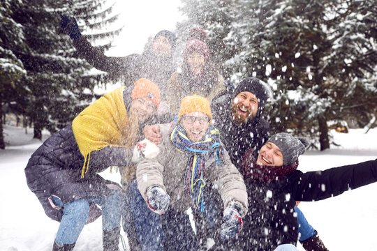Group of happy millennial friends having fun in the snow. Bunch of cheerful adult people in warm hats, coats and jackets throwing snow in the air for a funny group photo in a winter forest