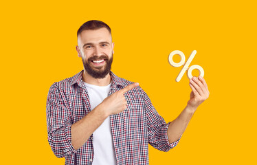 Studio portrait of smiling man with percentage sign. Happy young man in casual shirt isolated on orange yellow background advertising sale and pointing at discount percent symbol in his hand