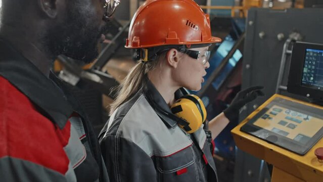 Caucasian female plant worker explaining African American male colleague how to operate CNC machine using special digital equipment with sensor screen
