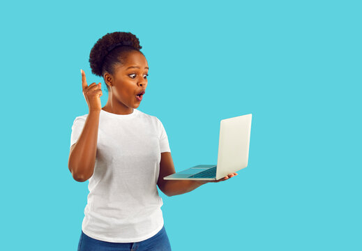Surprised african american woman with ponytale in white t-shirt having an idea looking at screen of laptop on blue background. Showing index finger up. Creating new creative ideas concept.