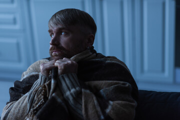 upset and frozen man looking away while sitting under warm blanket during energy shutdown.