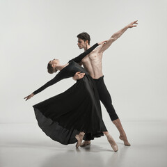Young muscular man and woman in black silk dress, ballet dancers performing isolated over grey studio background