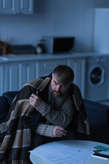 upset and frozen man sitting under warm blanket near invoices and money in twilight.