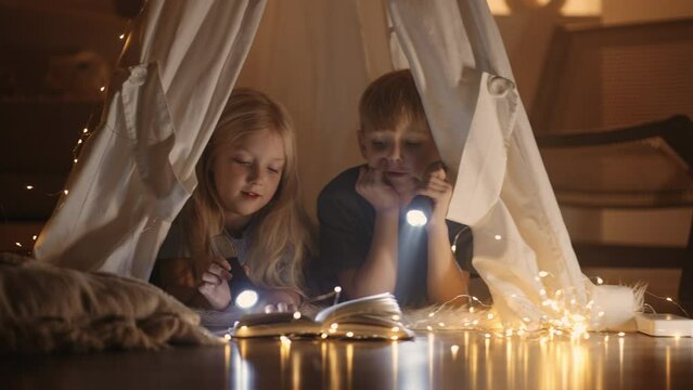 two little dreamers are reading book in wigwam in playroom, girl and boy play at home in evening