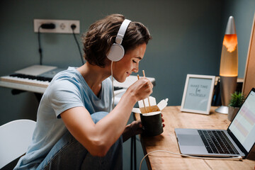 Young woman in headphones using laptop while eating asian food on bed