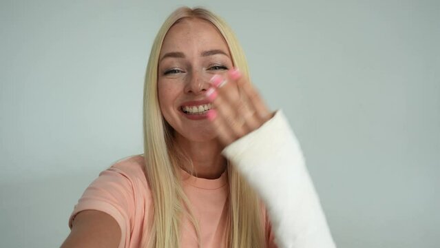 POV portrait of cheerful young woman with broken arm wrapped in white plaster bandage talking via video chat using mobile phone looking at camera, on white isolated background in studio, slow motion.