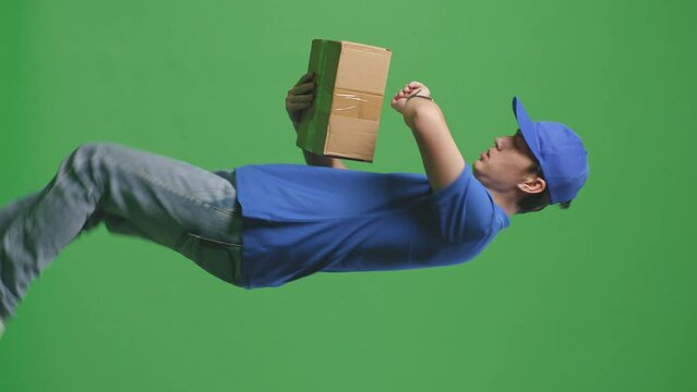 Side View Of Asian Male Courier In Blue Uniform Checking Watch And Running While Delivering A Carton On Green Screen Background In The Studio
