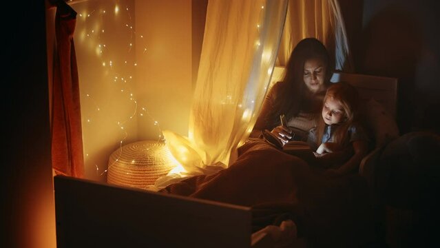 cute child girl and her mother are reading book together in dark bedroom in Christmas holidays