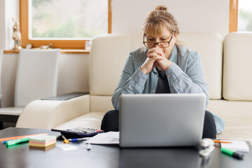 Sad caucasian middle aged female freelancer, tired of working overtime online, sitting at home in...