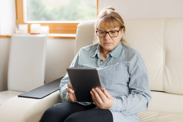 Beautiful mature woman in eyeglasses is using a digital tablet and smiling while sitting on couch at home