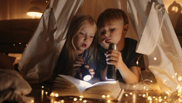 caucasian children are playing at home in night, boy and girl lying in teepee tent and reading book
