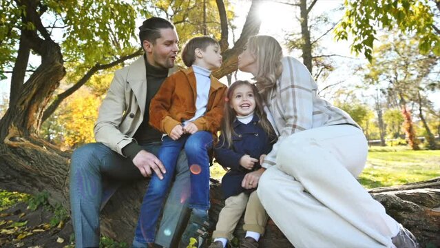 Happy family in an autumn park. Mother, father, son and daughter sitting on a tree trunk, mom kissing her son, yellowed trees around. Slow motion