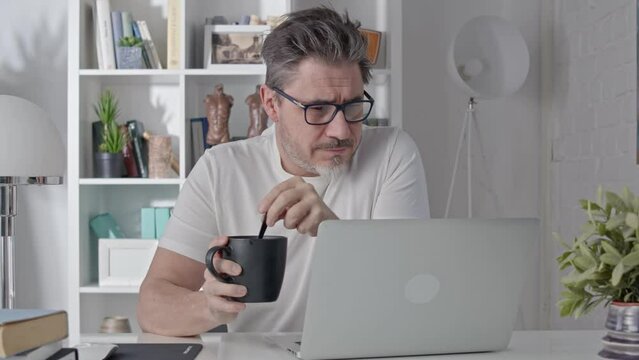 Middle aged man sitting at desk in morning at home, drinking coffee reading news on laptop. Businessman working in home office with computer, smiling.
