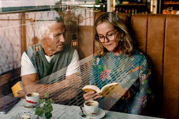Senior woman reading book to male friend sitting with arms crossed in cafe seen through glass