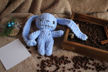 The blue hare lies on a table on a plush blanket. There is a wish card next to it. Coffee beans and a wooden tray complete the decor. Handmade. Hobby. Knitting. Gifts for the holidays.