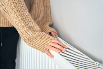 Child wearing brown woolly jacket warming cold hand in front of heating radiator in winter time. Electric or gas heater at home. Heating season, cold room, heating problems