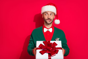Photo of doubtful unsure guy dressed xmas green cardigan bowtie looking empty space holding noel gift isolated red color background
