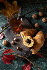 Cozy tea time: a glass on hot tea and chocolate croissants on a wooden board around by pumpkin, autumn leaves, nuts. Close up. Top view - 545196176