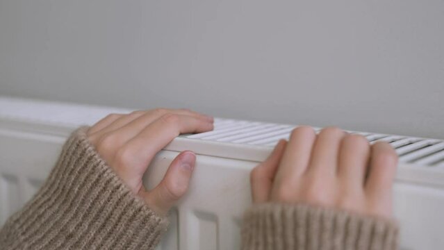 Girl wear woolly brown sweater warming cold hands in front of heating radiator in winter time. Electric or gas heater at home. The symbolic image of the heating season. Selective focus