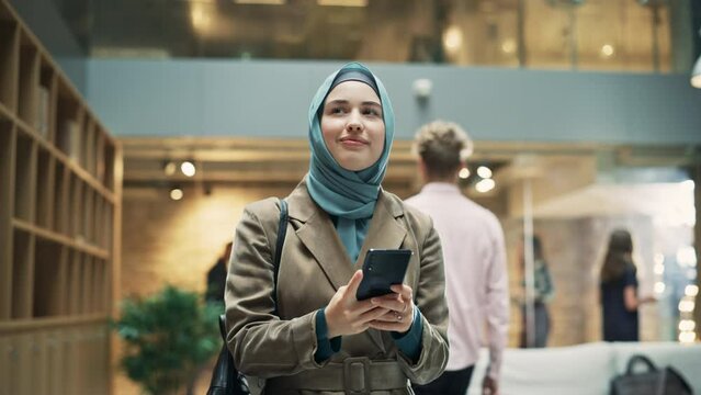 Portrait of Businesswoman Wearing Hijab Using Smartphone App to Share Social Media Post about Career Growth. Professional Female Manager Walking Through Office Building, Smiling and Looking Happy.