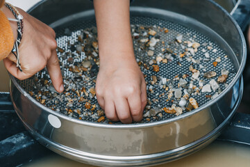 Child hand picking pebbles at the sieve at archaeological excavations or extraction of gold and other gems at the prospecting site. Muddy water in background. Hobby and recreation - obrazy, fototapety, plakaty