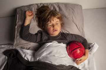 Healthy child, sweetest blonde toddler boy sleeping in a football ball. Dream of becoming a football player. Young soccer player. Beautiful baby boy have a healthy sleep in the bed. Sleeping baby.