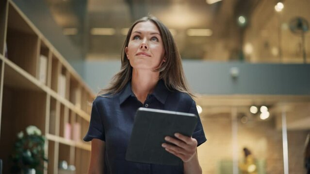 Close Up of a Content Female CEO Making Notes on her Tablet. Portrait of a Caucasian Woman Crossing a Corporate Office Hall, Smiling and Looking Confident. Low Angle, Slow Motion
