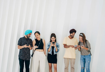 Portrait Of Smiling Friends Using Their Mobile Phones And Laughing While Standing In A Row Together In Front Of White Geometric Wall