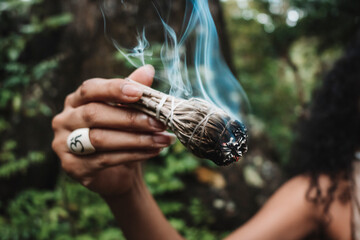 Imagen horizontal  de la mano de una mujer morena irreconocible sosteniendo un rollo de hojas de salvia encendido mientras hace un ritual.  