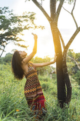 Imagen vertical de una mujer afroamericana de pie al lado de un árbol al aire libre realizando un ritual de purificación con rollo de hojas de salvia en sus manos. 