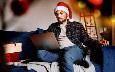 Alone sad man in Santa hat sitting on sofa using laptop and drink wine.