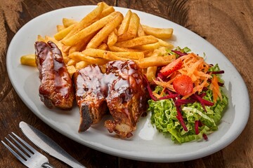 Top view of pork ribs with fries and vegetable salad in a white plate on a wooden table