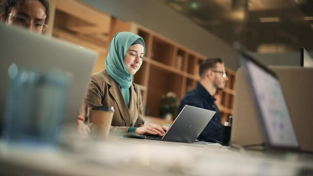 Portrait of Focused Muslim Woman Using Laptop in a Contemporary Spacious Office. Account Specialist Smiling While Having Productive Day. Active Colleagues Working on Computers in the Background.