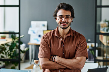 Young successful intern in eyeglasses and brown shirt keeping his arms crossed by chest while standing by workplace in front of camera - obrazy, fototapety, plakaty