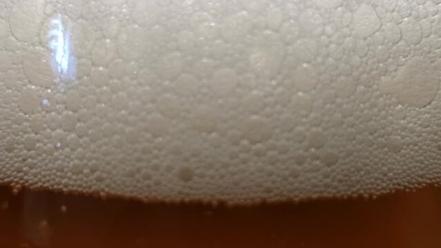 White thick beer foam. Fresh amber-colored beer is poured into a thin transparent glass glass. Above the liquid is a white dense patch with many bubbles.