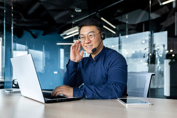 Cheerful and smiling office worker with video call headset, Asian customer support tech of online store, smiling and talking consulting. the man works inside the office.